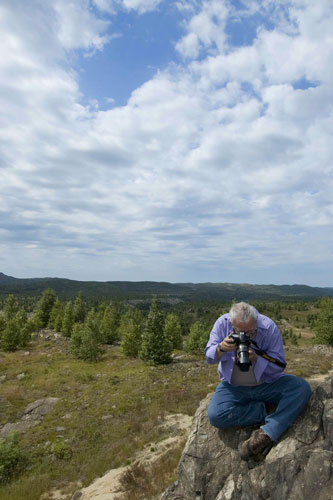 man holding camera facing down with lightly forested background a cloudy blue sky